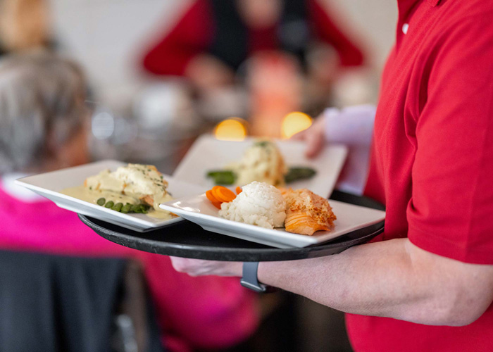 Server in a red shirt carrying a tray with plates of rice, asparagus, fish fillet, and vegetables, delivering meals to diners in a restaurant-style senior living dining room.