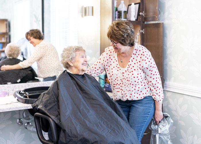 A senior resident smiles while sitting in a salon chair, covered with a black cape, as a hairstylist, wearing a white blouse with red hearts, chats with her in a friendly manner.
