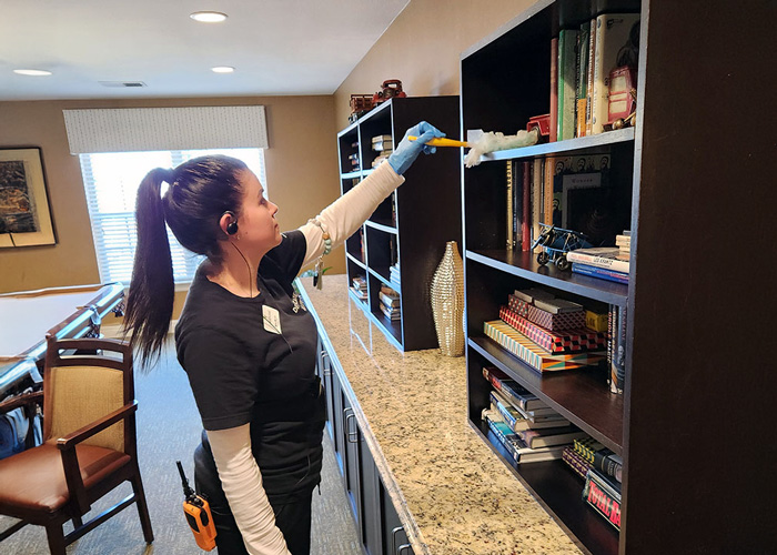 Staff member with a ponytail dusts a bookshelf in a senior living community, ensuring a clean and organized environment for senior residents to enjoy their activities.