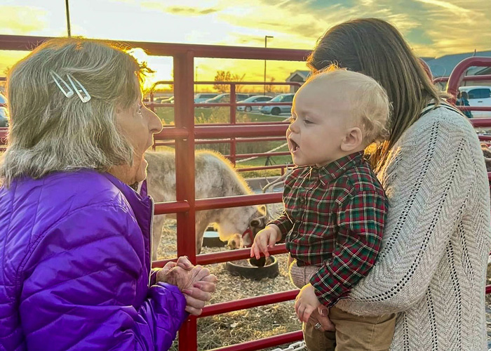 A senior resident in a purple jacket interacts joyfully with a baby held by a woman near a petting zoo, as the sun sets in the background, creating a warm and heartwarming moment.