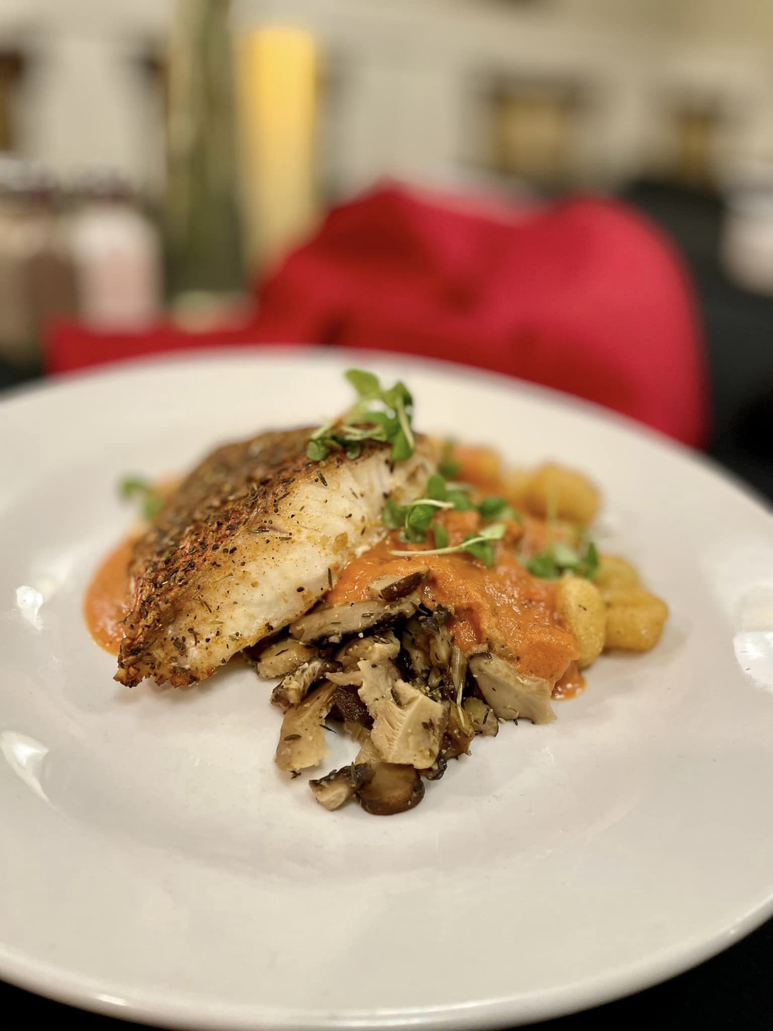 Seasoned fish fillet served with sautéed mushrooms, gnocchi, and a red pepper sauce, garnished with microgreens on a white plate, offering a gourmet dining experience.