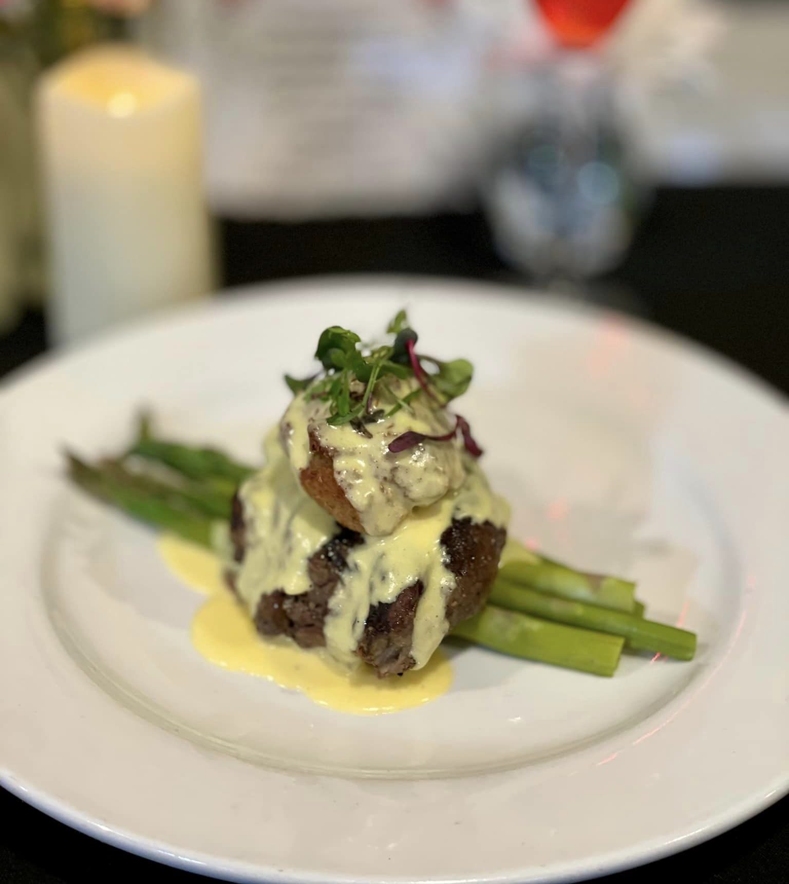 Plated gourmet steak with asparagus, topped with creamy hollandaise sauce and garnished with microgreens, set against a blurred background of a candle, flowers, and drink.