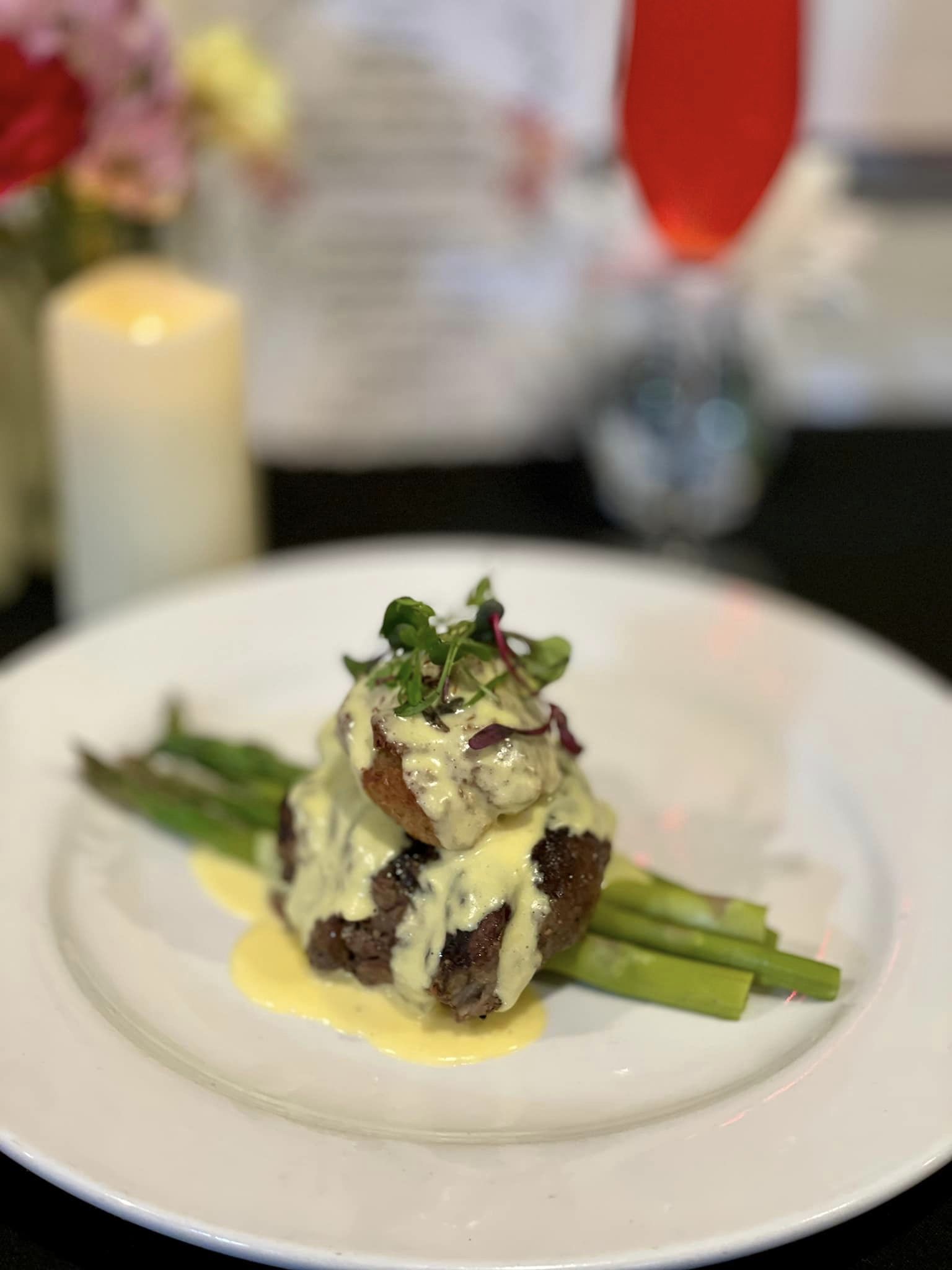 Plated gourmet steak with asparagus, topped with creamy hollandaise sauce and garnished with microgreens, set against a blurred background of a candle, flowers, and drink.