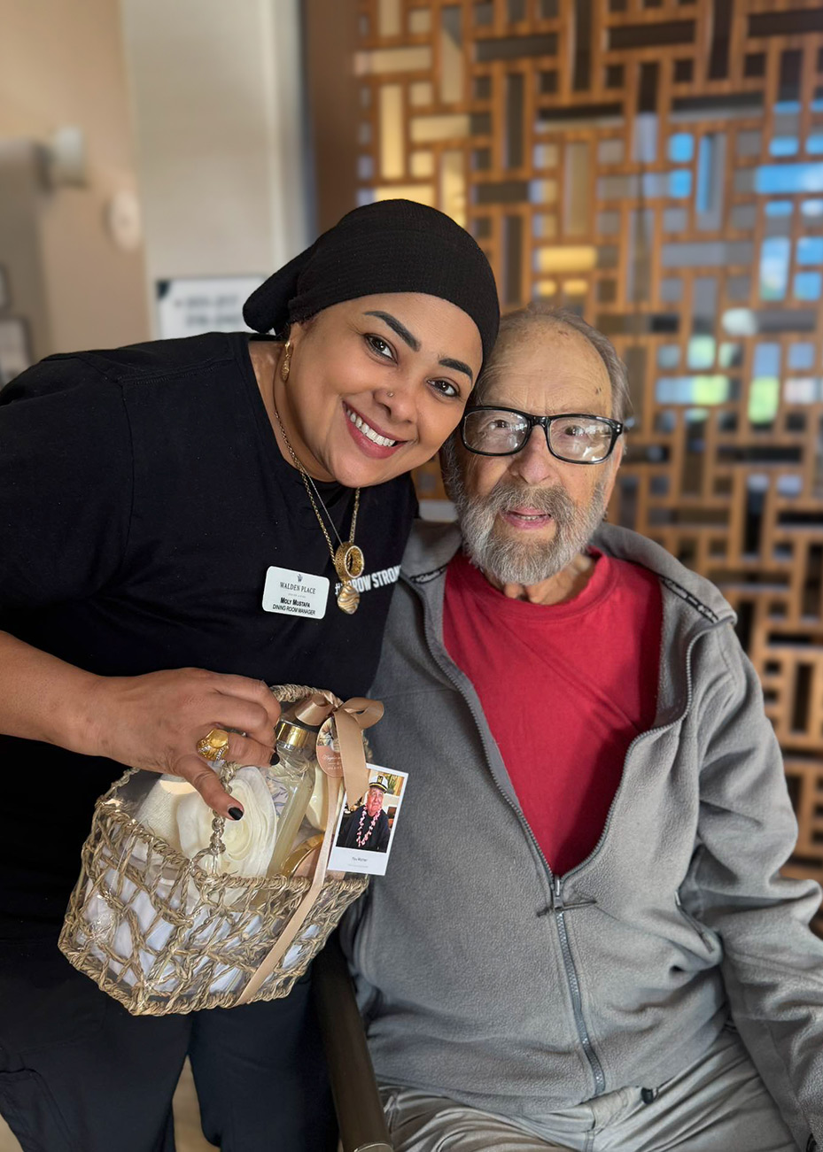 A smiling team member holds a gift basket alongside a senior man in a gray jacket.