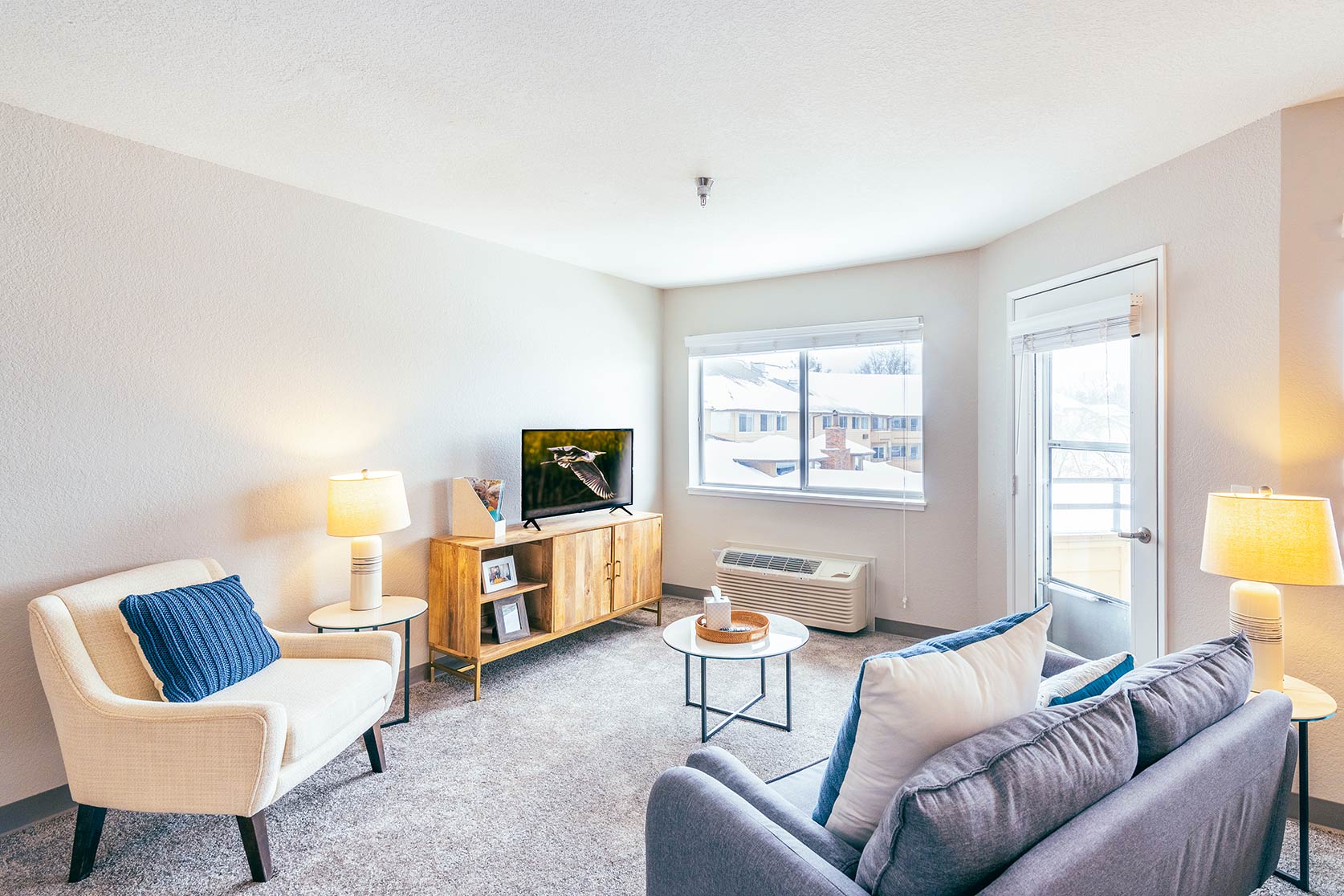 Bright senior living apartment living room with sofa, armchair, TV console, soft lighting, and large window overlooking snowy neighborhood.