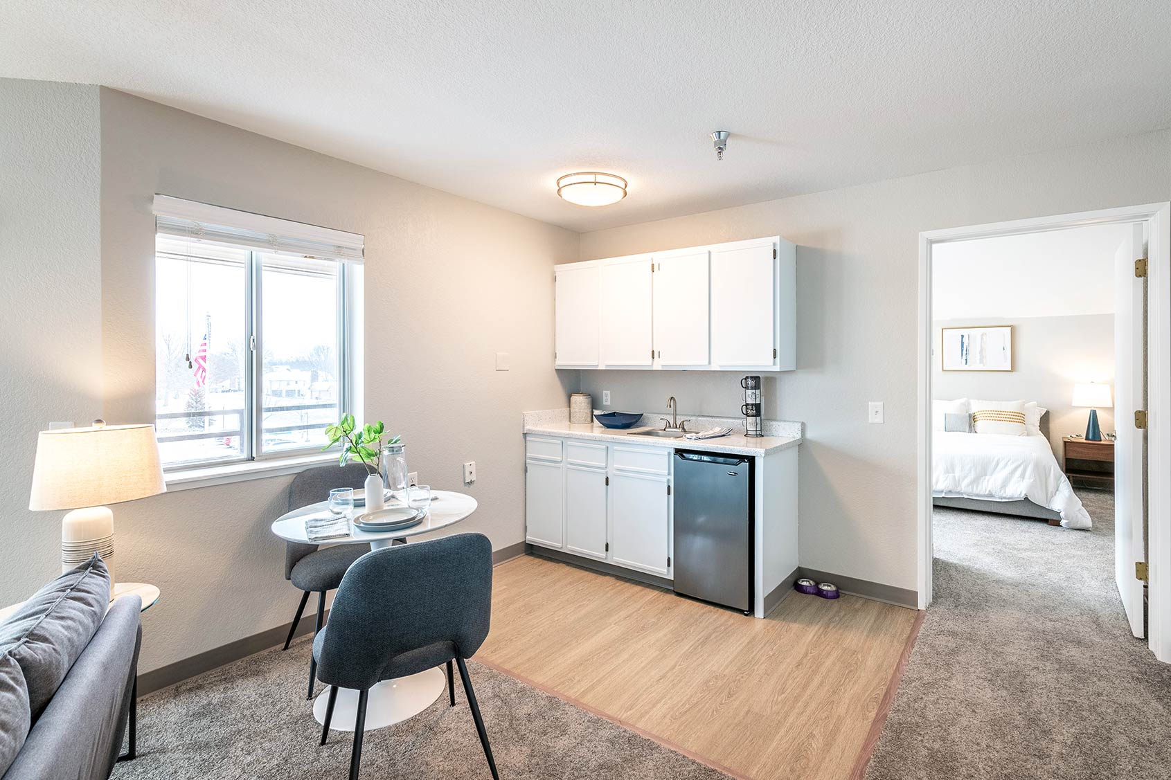 Senior living apartment kitchenette with white cabinets, mini refrigerator, small dining table for two, and open doorway to bedroom.