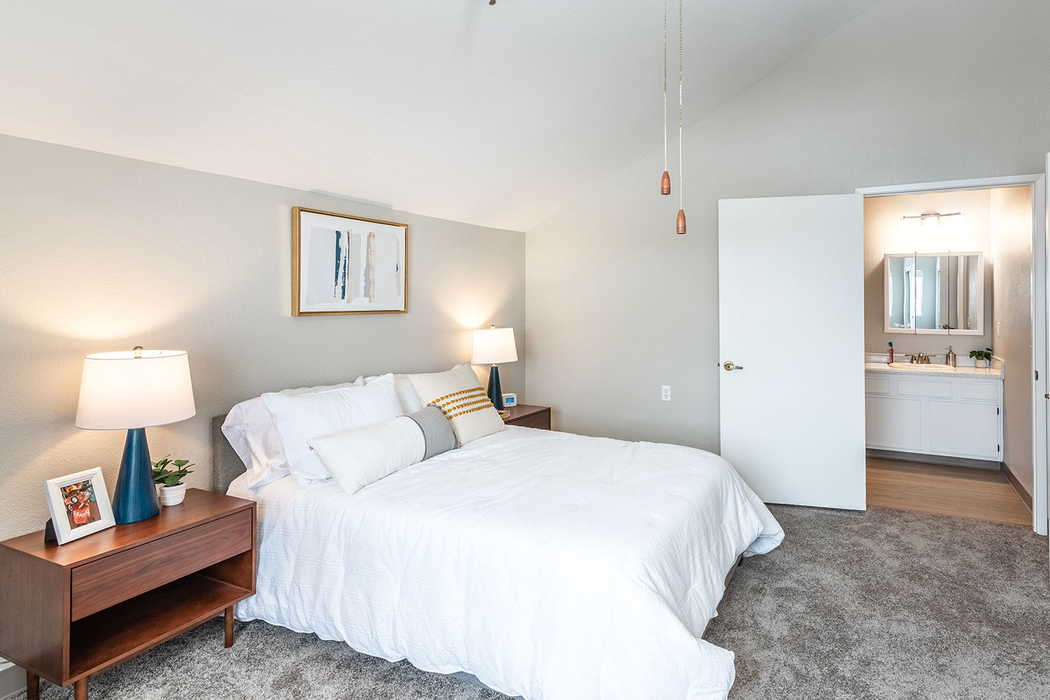 Spacious senior living bedroom with large bed, nightstands, soft lighting, neutral décor, and attached bathroom entrance.