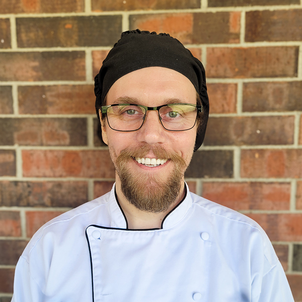 Brian Phillippe, Culinary Director at Buckingham Senior Living, smiling in a professional headshot, wearing a white chef coat, black chef hat, and glasses, standing in front of a brick wall background.