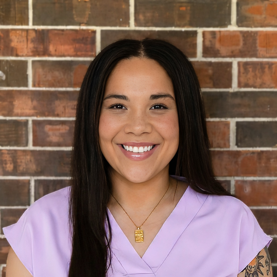 Maleah Hess, Senior Living Director at Buckingham Senior Living, smiling in a professional headshot with long straight dark hair, a lavender blouse, a gold pendant necklace, and a red brick wall background.
