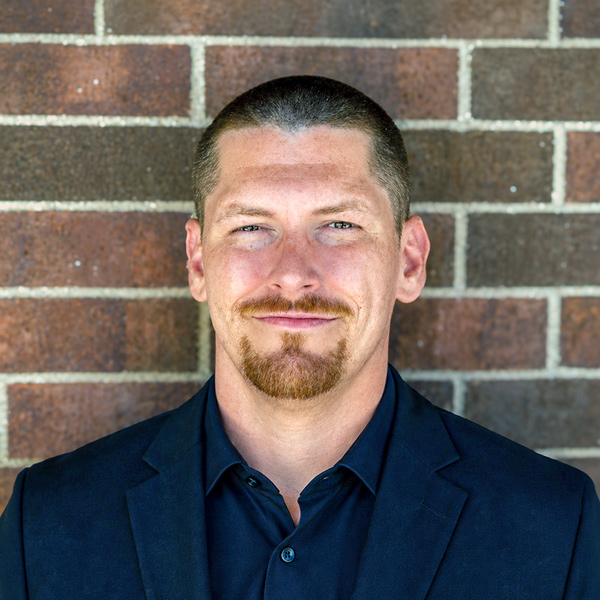 Aaron Thompson, Plant Operations Director at The Buckingham Senior Living, looking at the camera in a professional headshot with closely cropped hair, a goatee, a navy blazer, and a brick wall background.