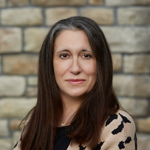 Amy Steinbeck, Senior Living Director at The Parkway Senior Living providing Interim Executive Director support at The Buckingham Senior Living, looking at the camera in a professional headshot with long dark hair, a patterned cardigan, and a stacked stone wall background.